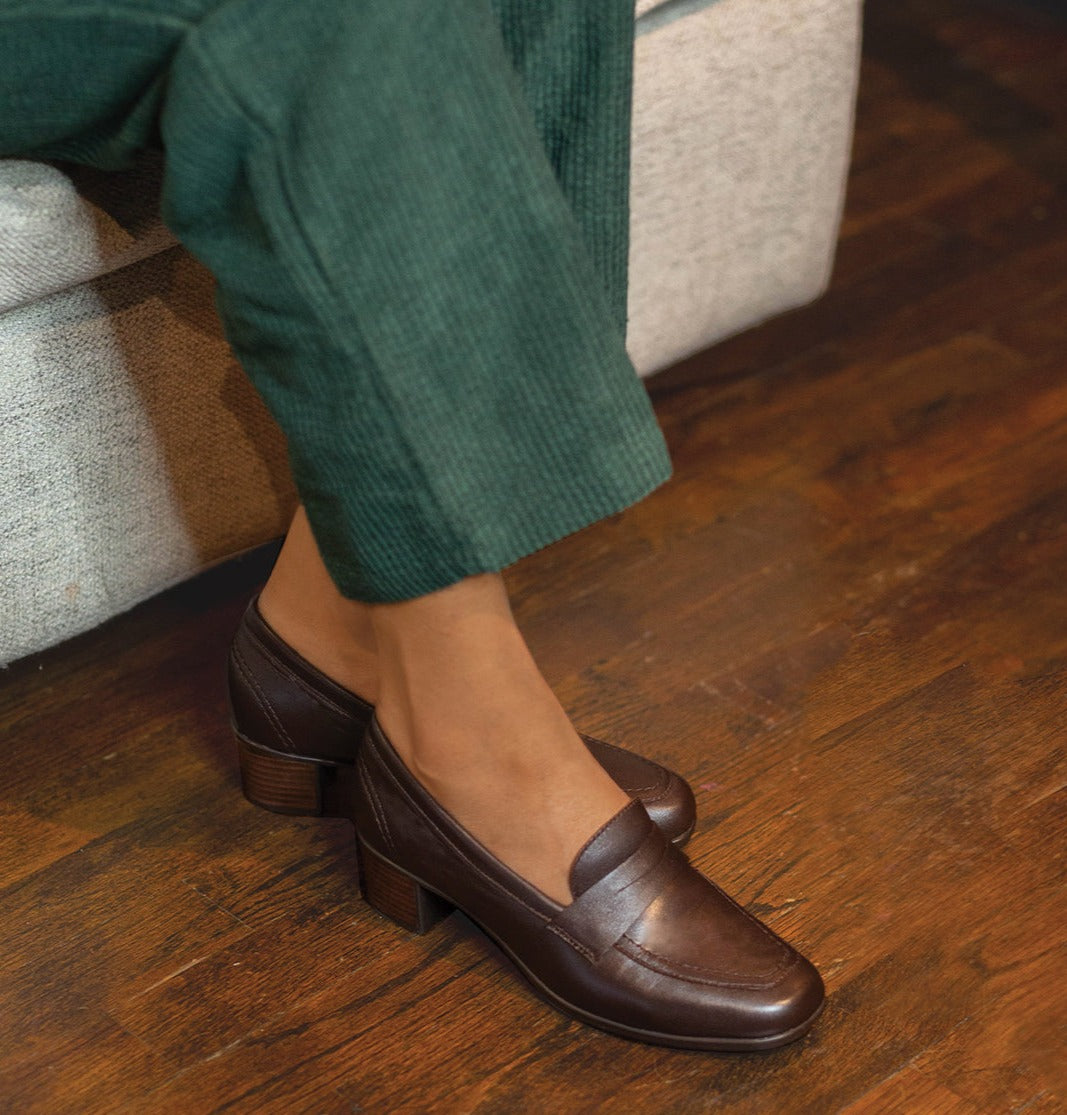 MUNRO CHOCOLATE COLOR LEATHER PENNY LOAFER PUMP ON MODEL WEARING GREEN CORDUROY SLACKS SITTING ON GRAY OTTOMAN-LIFESTYLE