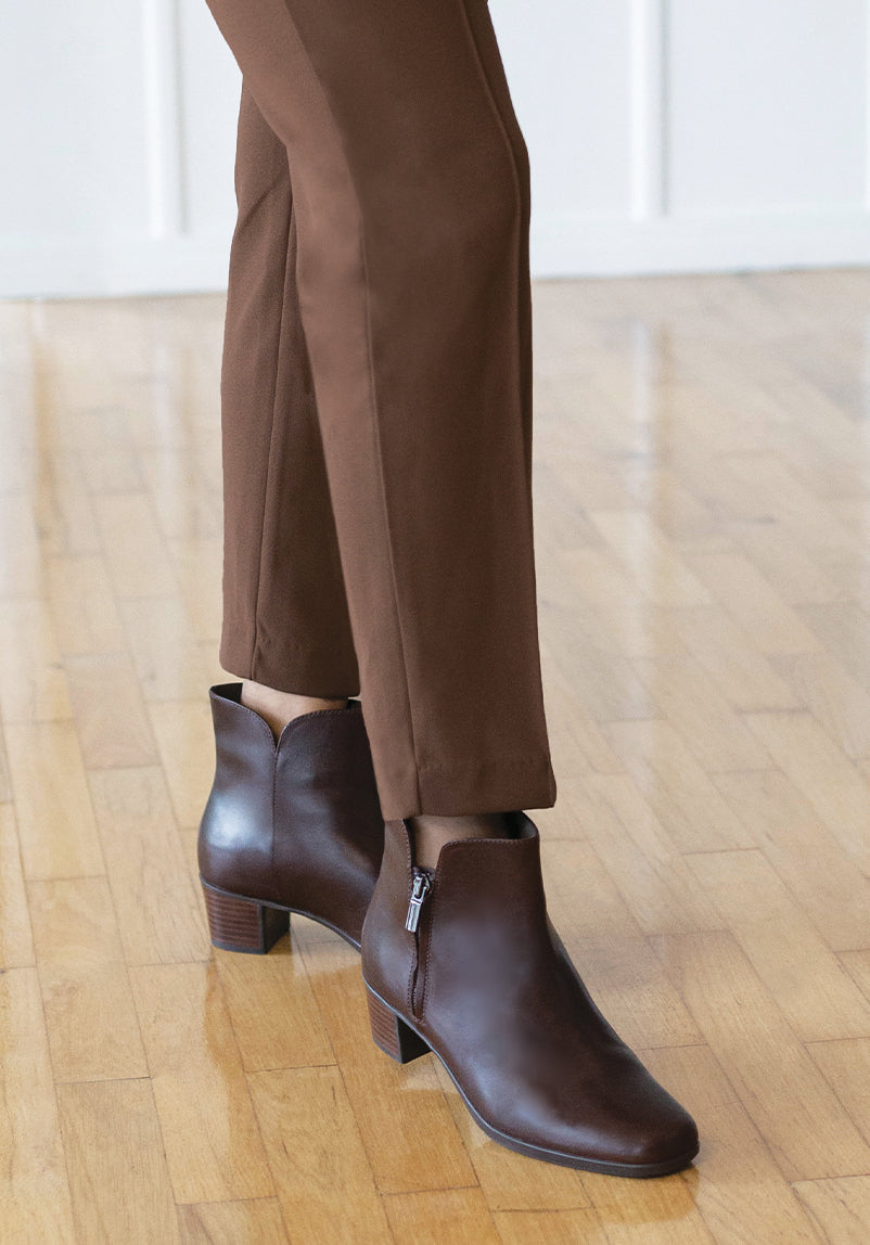 MUNRO LANDYN ANKLE BOOT IN FUDGE COLOR LEATHER WITH ZIPPER ON OUTSIDE ON MODEL WEARING BROWN SLACKS STANDING-LIFESTYLE